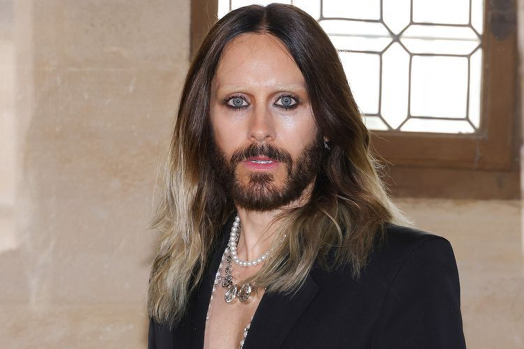 Auto-generated description: A person with long, wavy hair, a beard, and striking eye makeup is wearing a black blazer and layered necklaces while standing indoors near a window with a geometric design.